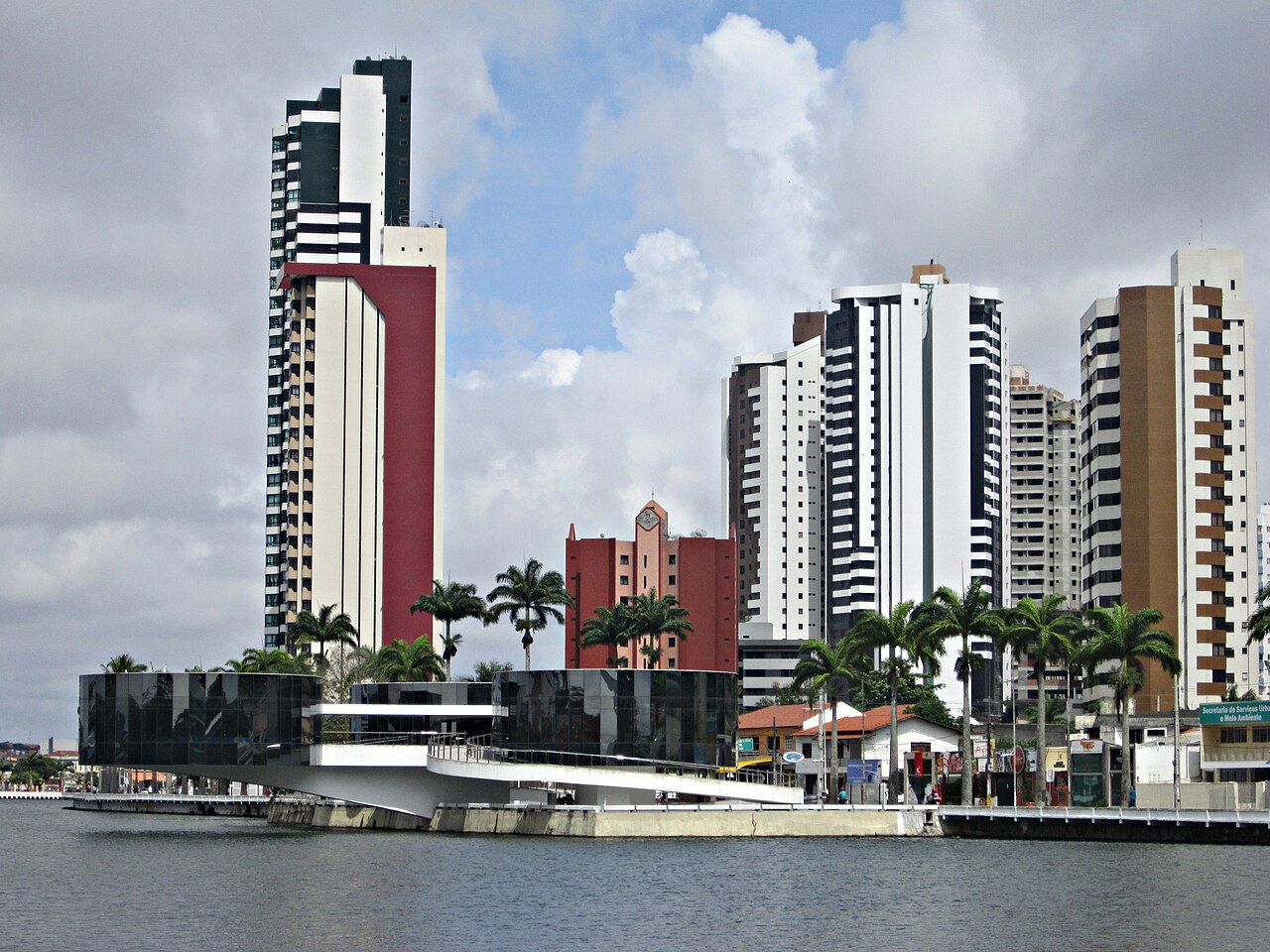 Açude Velho e Museu de Arte Popular da Paraíba em Campina Grande, com prédios altos e palmeiras sob céu com nuvens